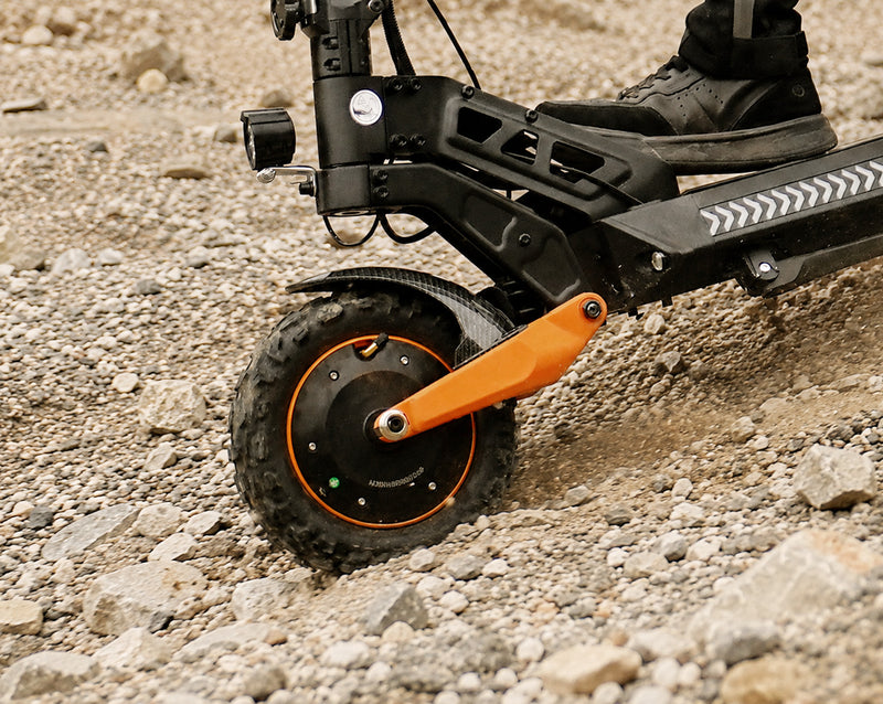 Close-up of an off-road electric scooter's front wheel, knobby tire, and orange suspension swingarm on a rocky trail.
