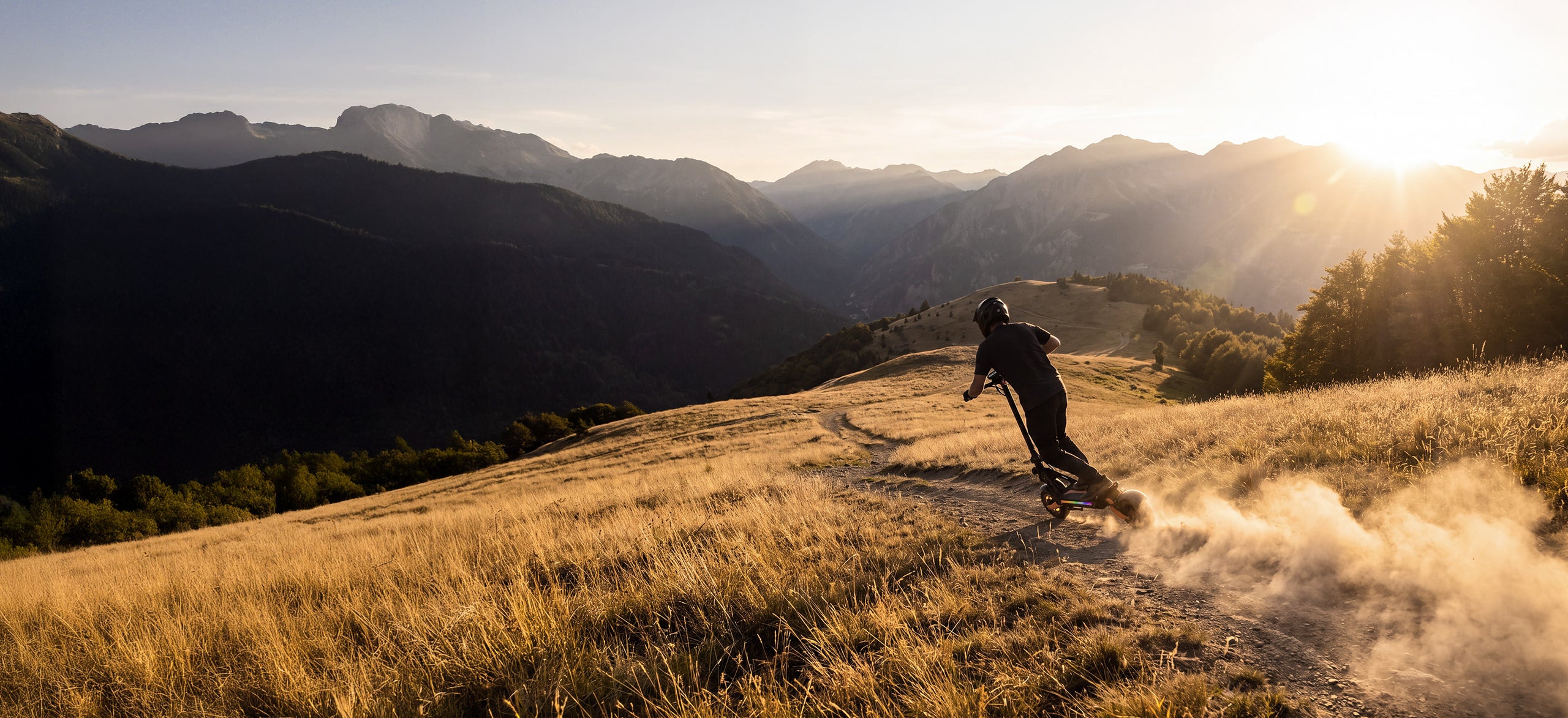 A person riding an off-road electric scooter down a dirt path at sunset with mountain scenery.
